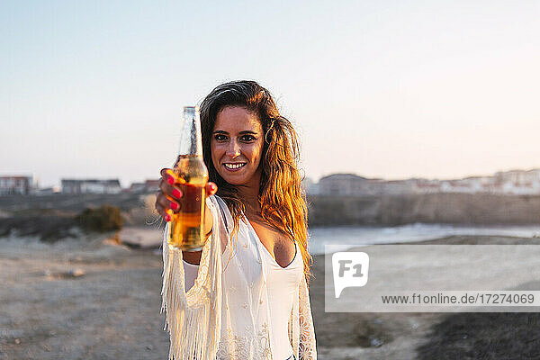 Lächelnde Frau zeigt kleine Weißweinflasche am Strand bei Sonnenuntergang