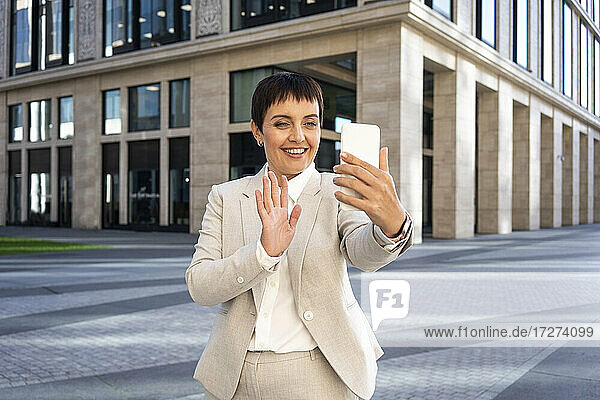 Businesswoman waving hand to video call while standing against modern office building