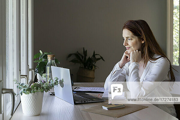 Weiblicher Arzt mit Hand am Kinn  der zu Hause sitzend einen Videoanruf auf dem Laptop hört