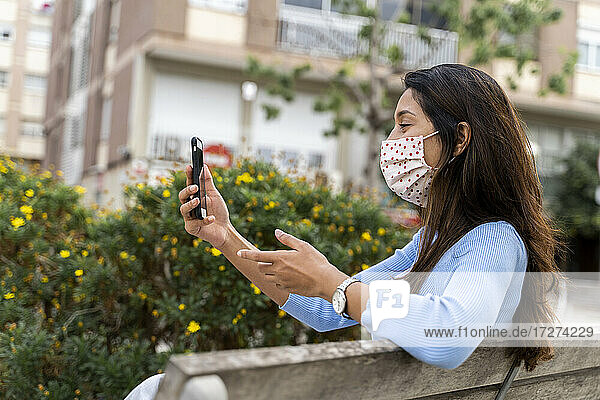 Frau gestikuliert beim Benutzen eines Smartphones auf einer Bank in der Stadt während COVID-19