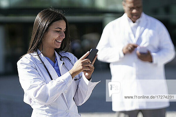Lächelnde Frau  die ein Mobiltelefon benutzt  während sie mit einem Arzt im Hintergrund vor einem Krankenhaus steht