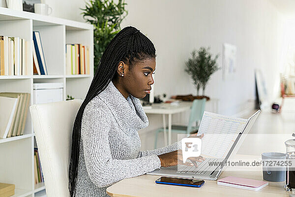 Female student concentrating while reading paper studying on laptop at home