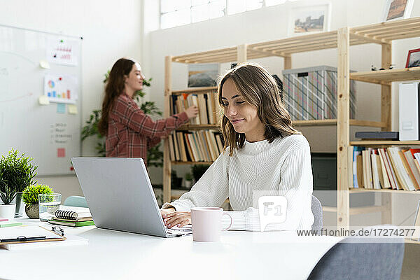 Lächelnde Geschäftsfrau  die an einem Laptop arbeitet  während ein Kollege im Hintergrund im Büro steht