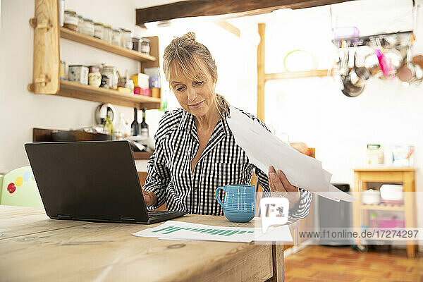 Ältere Frau arbeitet an einem Laptop und hält Papier in der Hand  während sie in der Küche zu Hause sitzt