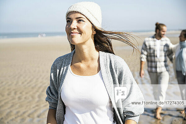 Lächelnde Frau blickt auf  während sie mit ihrem Freund und ihrer Mutter im Hintergrund am Strand spazieren geht