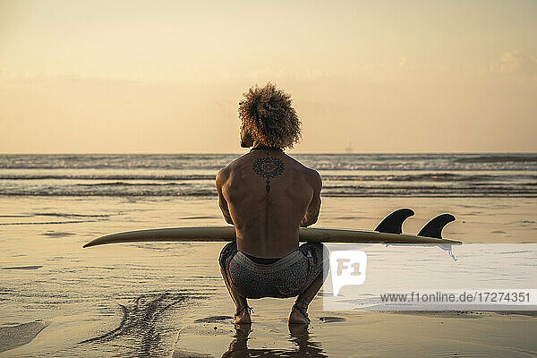 Junger Mann mit Surfbrett bei Sonnenuntergang am Meer hockend