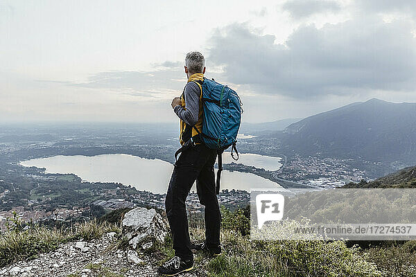 Mature man with backpack looking at lake and city against sky during sunset