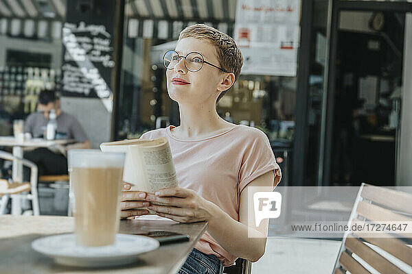 Mittlere erwachsene Frau  die mit einem Buch wegschaut  sitzt in einem Straßencafé an einem sonnigen Tag