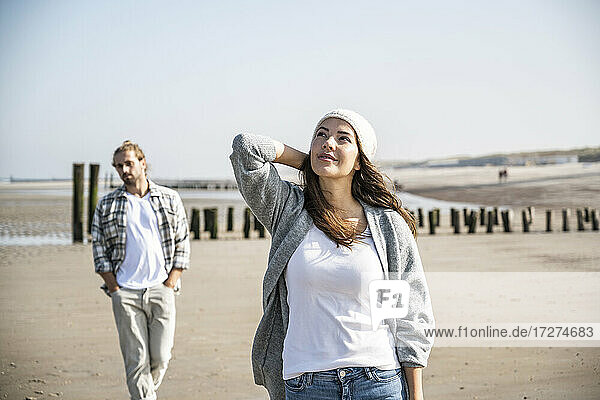 Thoughtful young woman looking up while man walking in background at beach