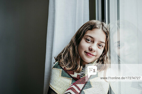 Smiling girl covered in blanket sitting by window at home