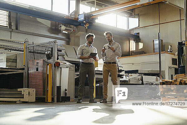 Businessman with digital tablet standing by colleague checking metal object at factory