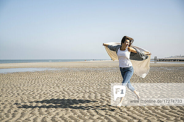 Carefree young woman running while holding blanket at beach against clear sky