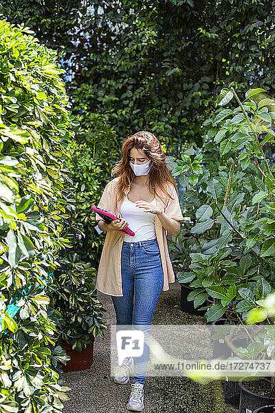 Geschäftsfrau mit Gesichtsmaske  die ein digitales Tablet benutzt  während sie in einem Gartencenter spazieren geht