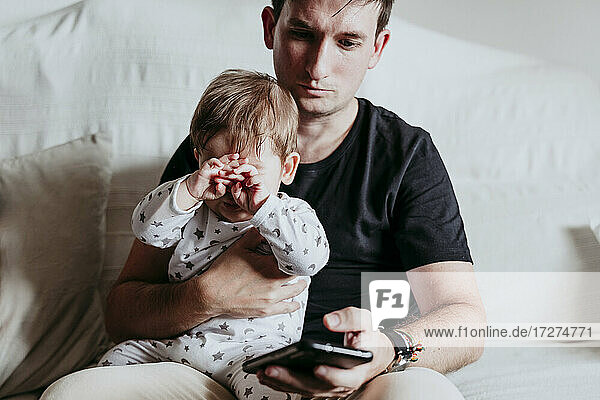 Vater benutzt sein Smartphone  während er mit seinem Sohn zu Hause auf dem Sofa sitzt