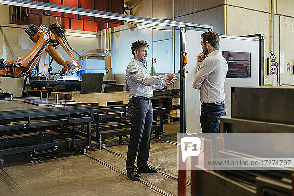 Junger und reifer Geschäftsmann diskutieren  während sie in einer Fabrik vor einer Robotermaschine stehen