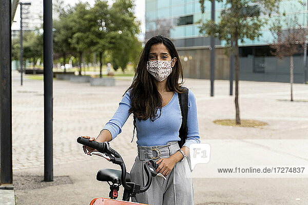 Beautiful woman wearing protective face mask while standing with bicycle in city during COVID-19 pandemic