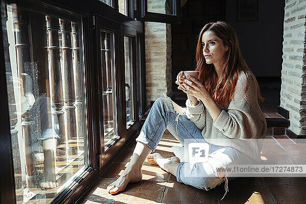 Junge Frau trinkt Kaffee  während sie zu Hause auf dem Boden sitzt