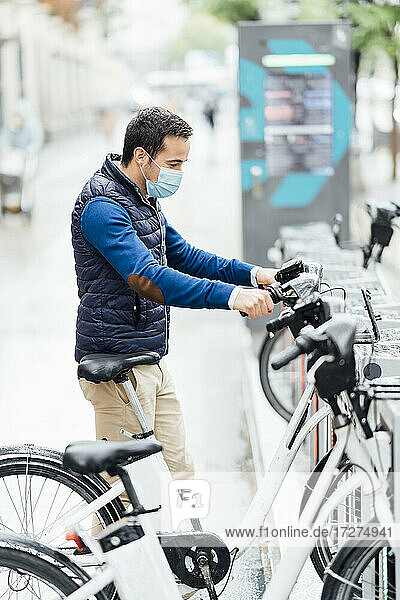 Junger Mann mit Gesichtsschutz  der ein Elektrofahrrad an einer Parkstation während COVID-19 hält
