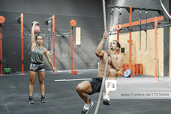 Young man exercising with rope while girlfriend lifting kettlebell in gym