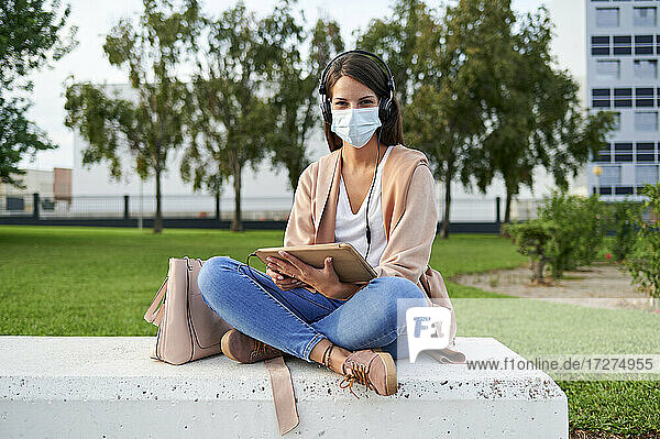 Junge Frau  die ein digitales Tablet benutzt und eine Gesichtsmaske trägt  während sie auf einer Bank in einem öffentlichen Park sitzt