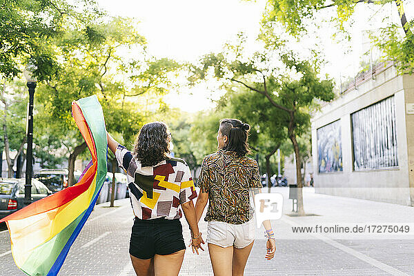 Lesbisches Paar mit Regenbogenfahne hält sich auf dem Fußweg an den Händen