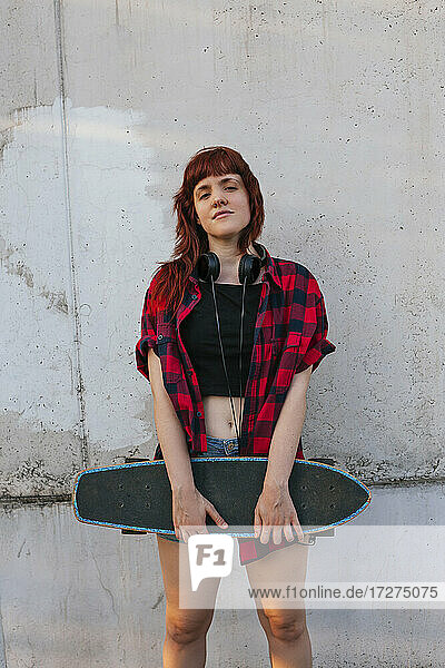 Junge rothaarige Frau mit Kopfhörern hält Skateboard gegen die Wand