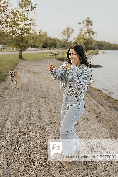 Smiling woman dancing while standing with dog running in background by lake