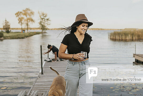 Lächelnde Frau hält ein Weinglas  während sie mit einem Hund und einem Mann im Hintergrund auf einem Pier spazieren geht