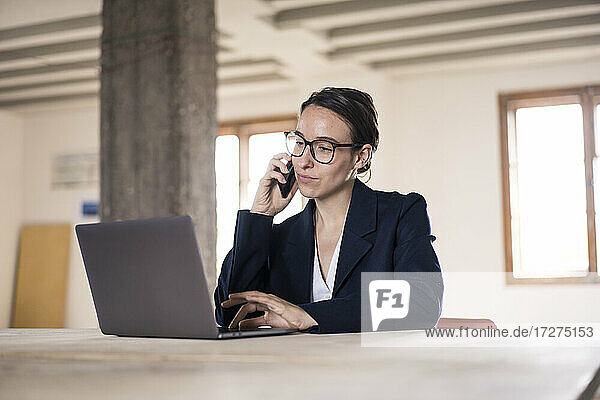 Businesswoman talking on mobile phone while working on laptop at office