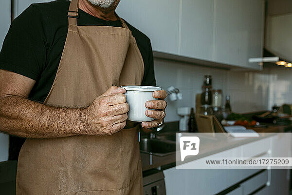 Mittelteil eines Mannes  der eine Kaffeetasse hält  stehend in der Küche zu Hause