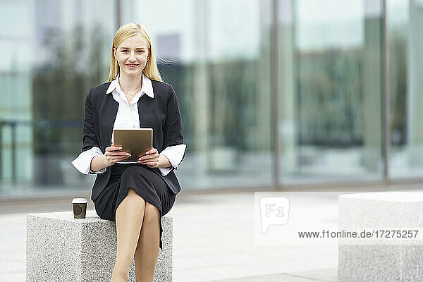 Lächelnde Geschäftsfrau  die ein digitales Tablet benutzt  während sie auf einer Bank vor einem Gebäude sitzt