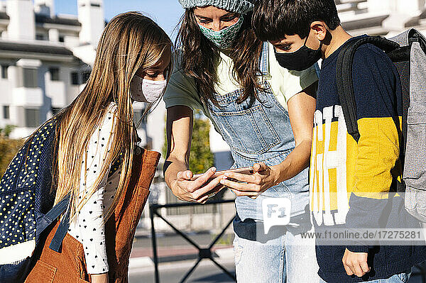 Mother and kids wearing protective face mask using smart phone standing in public park on sunny day