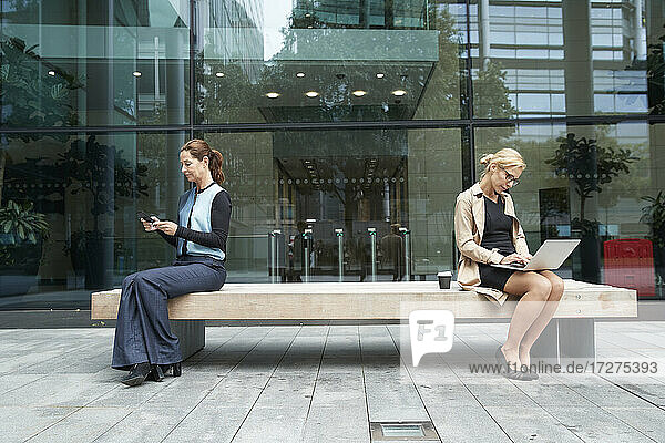 Geschäftsfrau und Kollege arbeiten im Sitzen mit sozialem Abstand auf einer Bank vor einem Bürogebäude