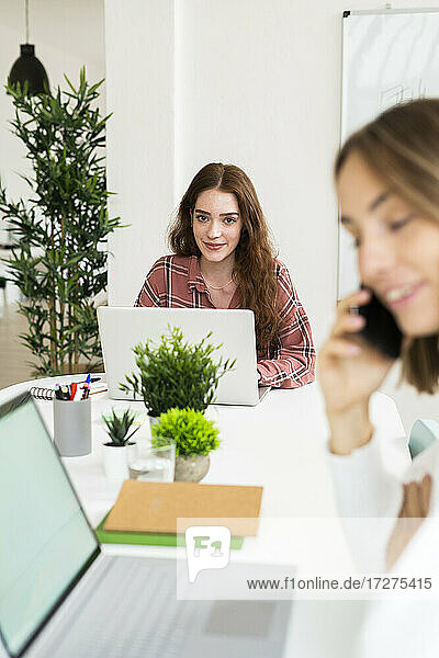 Lächelnde junge Frau  die an einem Laptop arbeitet  mit einer Geschäftsfrau  die im Vordergrund mit einem Mobiltelefon spricht  im Büro