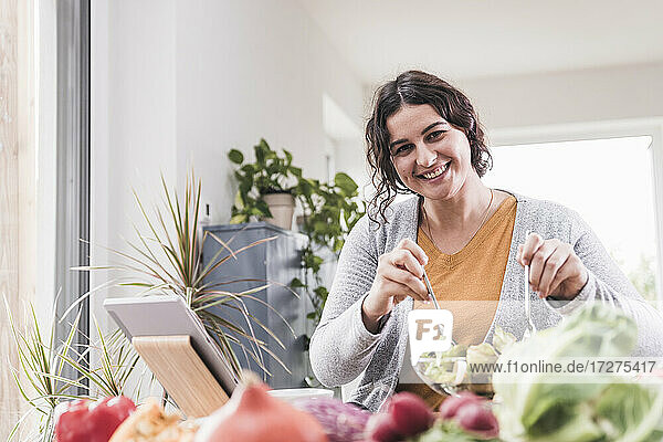 Lächelnde Frau  die Salat isst  während sie ein digitales Tablet zu Hause benutzt