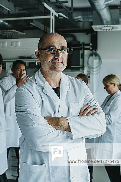 Lächelnder männlicher Wissenschaftler mit verschränkten Armen  der mit einem Mitarbeiter im Hintergrund in einem Labor steht