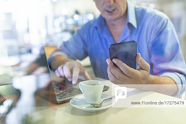 Geschäftsmann hält sein Smartphone in der Hand und benutzt seinen Laptop in einem Café