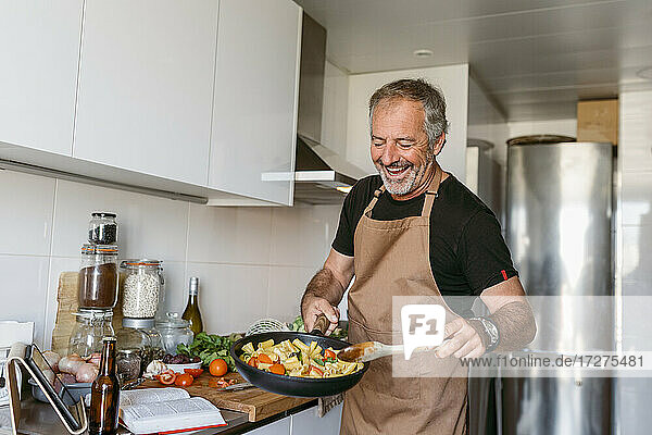 Lächelnder reifer Mann bereitet Nudeln zu  während er in der Küche zu Hause steht