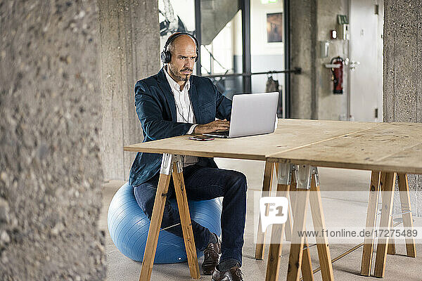 Geschäftsmann mit Kopfhörern  der einen Laptop benutzt  während er auf einem Fitnessball im Büro sitzt