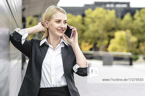 Lächelnde Geschäftsfrau  die wegschaut  während sie mit einem Handy in der Stadt telefoniert