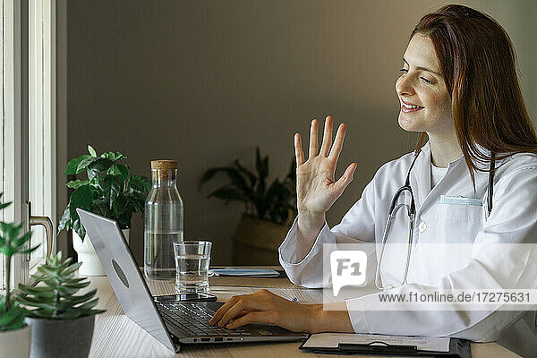 Junge Ärztin winkt dem Patienten zu  während sie eine Online-Konsultation von zu Hause aus durchführt