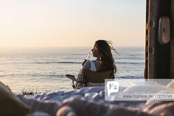 Junge Frau trinkt Weißwein  während sie auf einem Stuhl neben einem Wohnmobil am Strand bei Sonnenuntergang sitzt