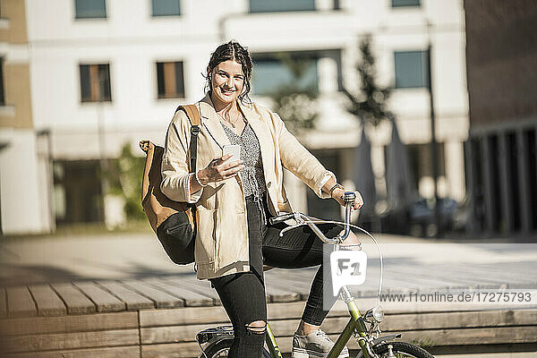 Lächelnde Pendlerin mit Smartphone auf dem Fahrrad in der Stadt an einem sonnigen Tag