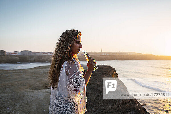 Junge Frau mit kleiner Weißweinflasche  die am Strand steht und wegschaut