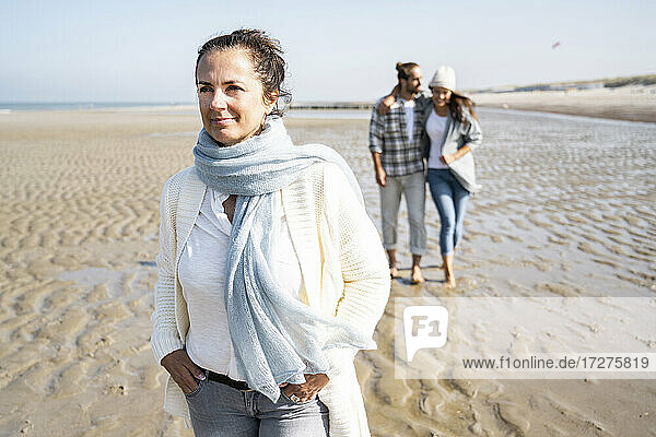 Frau mit Händen in den Taschen schaut weg  während sie mit einem Paar im Hintergrund am Strand spazieren geht