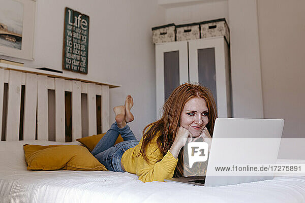 Smiling woman lying on front with hand on chin using laptop at home