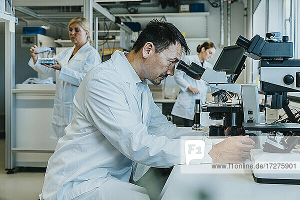 Männlicher Wissenschaftler sitzt am Mikroskop  während ein Mitarbeiter im Hintergrund im Labor arbeitet