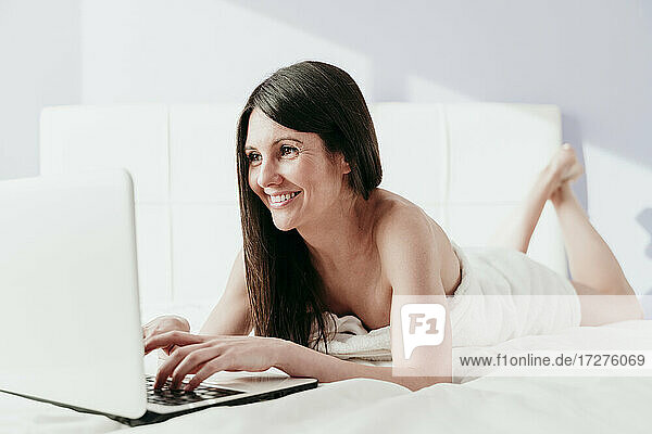 Lächelnde schöne Frau  die einen Laptop benutzt  während sie zu Hause auf dem Bett liegt