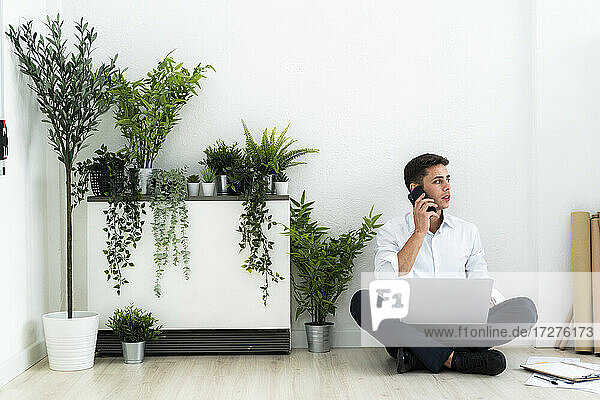 Businessman talking through mobile phone while sitting with laptop on floor against white wall in office