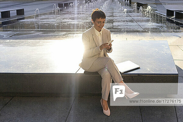 Geschäftsfrau  die ein Mobiltelefon benutzt  während sie an einem Springbrunnen in der Stadt sitzt